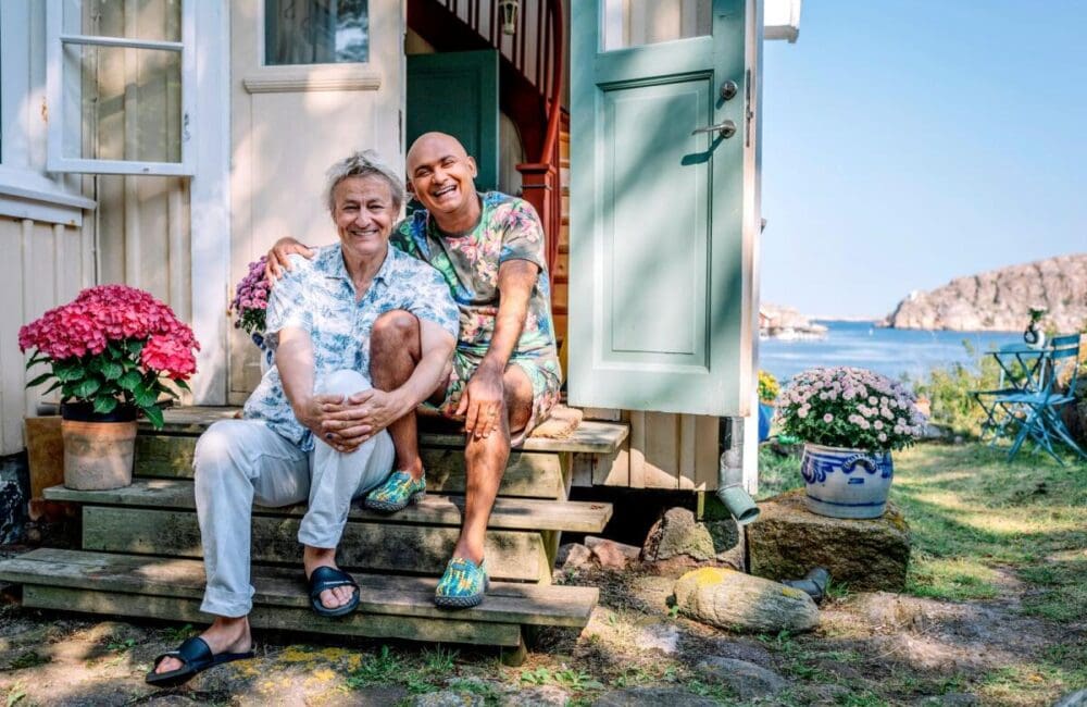 På en trappa framför ingången ett äldre trähus i ljusa pastellfärger, sitter Lars med ena knäet uppdraget, han omfamnar både sitt och Juniors knä. Junior sitter bredvid i med ena armen om Lars. De har ljusa, mönstrade och färgglada kläder på sig. I bakgrunden skymtar havet och Bohusläns klippor.