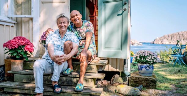 På en trappa framför ingången ett äldre trähus i ljusa pastellfärger, sitter Lars med ena knäet uppdraget, han omfamnar både sitt och Juniors knä. Junior sitter bredvid i med ena armen om Lars. De har ljusa, mönstrade och färgglada kläder på sig. I bakgrunden skymtar havet och Bohusläns klippor.