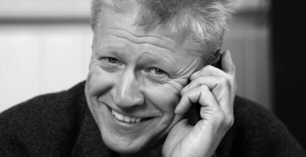Svartvitt porträtt på Martin som har en telefon tryckt mot örat och leende tittar in i kameran.